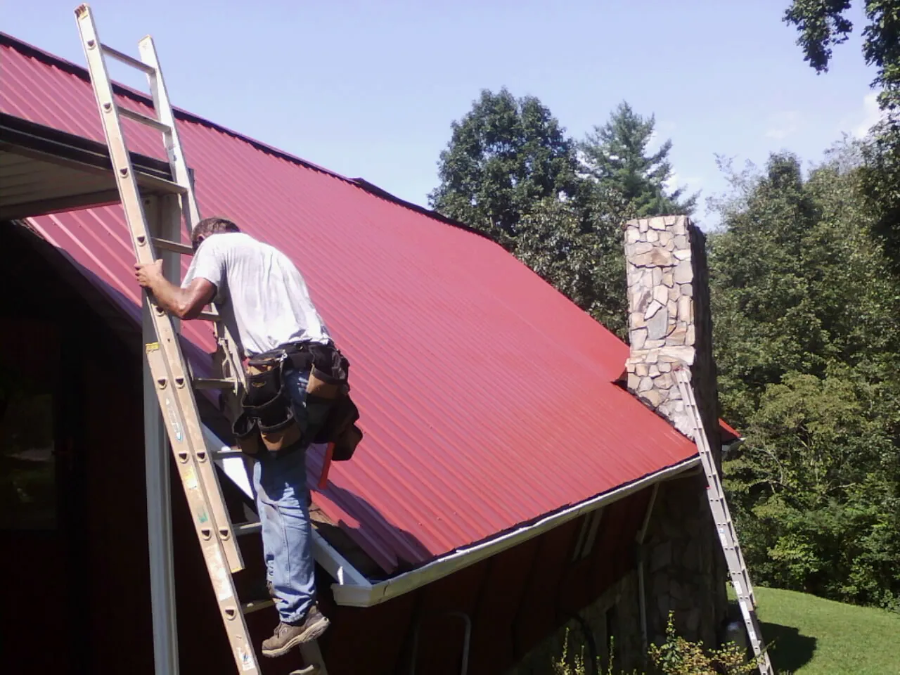 Roofer on a ladder beside a red metal roof and stone chimney — Soffit & Fascia Repair by Roofing Brothers in Zenia